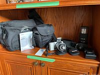 Full lot of camera kit including camera body, lenses, bags, manuals, charger, and battery displayed on wooden shelf under natural light.