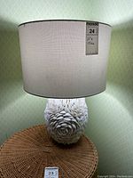 Full view of the table lamp showing the white fabric drum shade and the shell-covered base on a wicker surface against a patterned green wall.
