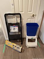 Wide view of all lot items against door: black aluminum ladder, black resin ladder, white plastic step stool, blue bin on stool, Easy Move sliders in box on floor.
