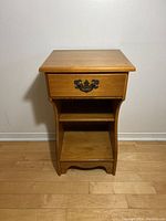 Front view of the maple nightstand showing drawer with metal handle and lower shelf, positioned on wooden floor against a white wall.