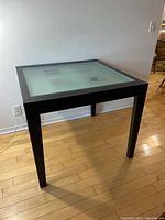 Square hardwood extendable table with frosted tempered glass top featuring clear square insets, black frame and legs, on wooden floor against plain wall.