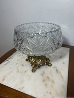 Side view of a clear crystal bowl with detailed starburst cut patterns, sitting on an ornate brass footed base, placed on a marble surface.