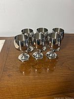 Set of six metal goblets arranged on a wooden surface, showing their rounded bowl shape and reflective exteriors.