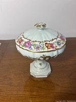 Side view of white porcelain powder box with domed lid, footed hexagonal base, hand painted pink and blue flowers and gold detailing.