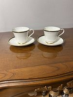 Two white porcelain cups and saucers with gold rim detailing on a wooden surface.