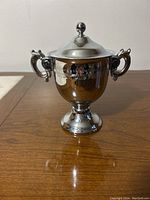 Front view of a shiny silver sugar bowl with two ornate handles, lid with a round knob, standing on a pedestal base.