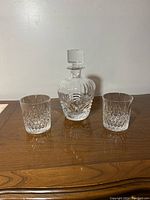 Full set including crystal decanter with stopper and two matching crystal glasses displayed on wooden surface against plain background.