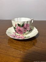 Side view of the teacup placed on its matching saucer, showing the pink rose floral pattern and scalloped edges.