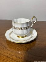 White porcelain teacup and saucer with gold chain design and gold handle on wood surface, side angle view