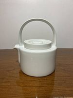 Front view of modernist white ceramic teapot with cylindrical body, handle, and spout on wooden surface.