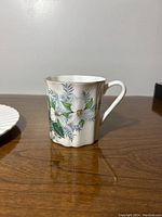 Fine bone china coffee mug with white Trillium flowers and green leaves, standing on wooden surface, photographed from different angles showing floral detail and handle