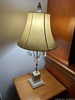 Full view of crystal table lamp with beige bell shade on wooden surface.