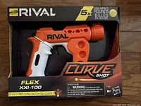 Front view of the Nerf Rival Curve Shot Flex XXI-100 blaster in original packaging, showing orange and white blaster and 5 yellow high-impact rounds.