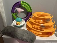 Photo of all cat toys grouped together including multi-level orange track toy, circular green and purple toy, feathered ball toy on spring, and long grey plush toy.