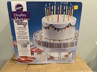 Front of the box showing the Wilton customizable cake pedestal with colorful striped edge on the top plate and the cake with candles.