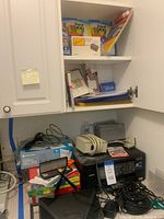 Overview of Brother printer, label maker, calculator, cables, and office supplies in cabinet and on shelf