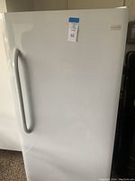 View of the front of a white upright freezer with handle and a hanging lot tag labeled '82'.
