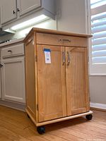 Wooden kitchen cart against kitchen cabinetry and window blinds, showing front and side view with closed doors and top drawer with metallic handle.