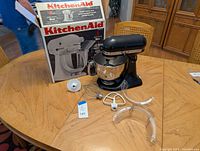 Black KitchenAid Artisan stand mixer on wooden table with original box, stainless steel mixing bowl, wire whip, flat beater, dough hook, and pouring shield accessories.