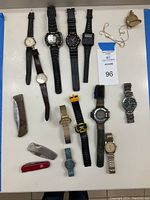 Photo showing multiple men’s wristwatches, pocket knives, and a pocket watch with chain laid out on a white surface for inventory.