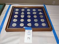 Top-down view of closed wooden case showing 25 silver medals beneath glass on blue velvet