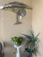 Decorative metal tree wall hanging mounted on beige stucco wall above the white pedestal plant stand holding a potted pothos plant, next to large succulent plants in gray pots on floor.