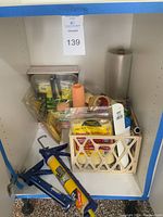 Photo of cabinet shelf containing blue caulk gun, orange foam paint rollers, masking tape, plastic paint grid, yellow plastic spreaders, and various packaged painting supplies.