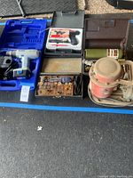 Top view of assorted power tools and accessories arranged on ground including cordless drill, hot glue gun, electric sander, sanding discs and various cases