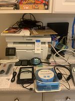 White HP printer with paper trays, black Kindle e-reader, Sony Walkman in packaging, white cables, calculators, and electronic accessories on countertop.