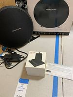 Harman/Kardon Onyx Studio portable speaker with original packaging box and power adapter, alongside boxed phone stand dock and warranty card laid out on surface.
