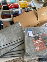 Folded gray tarp with grommets alongside packaged tarp on table with boxes and other items in background