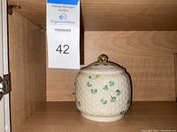 Vintage Belleek ceramic biscuit jar shown in wooden cabinet, displays cream color with textured basket weave and hand-painted green shamrocks.