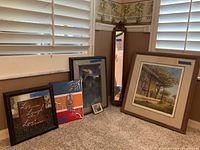 Photo showing five decorative wall items placed on carpeted floor near windows, including four framed prints and a full-length floor mirror with ornate top.