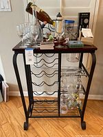 Front view of the wine rack with wine glasses on top, decanter, coasters, and decorative lighthouse bottle holder. Black metal frame with bottle slots and glass holders visible.