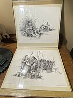 Two prints seen together on display; top print shows seated Indigenous figures by fire with pots, bottom print shows two figures in traditional clothing with hanging meat