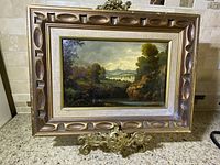 Antique landscape painting displayed on a decorative gold metal stand, showing full framed artwork on a tiled surface.