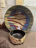 Front view of large ceramic bowl with abstract beige, red, and blue glaze and sandy textured rim. Smaller bowl placed in front shows cream textured exterior and black interior with irregular rim.