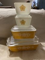 Five vintage Pyrex Butterfly Gold refrigerator storage dishes from 1970s stacked and arranged, showing yellow and white opaque glass containers, some with clear ribbed glass lids.