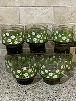 Front view showing 7 of the 8 vintage Libbey glasses with white daisies and green grass flower design against a tiled background.