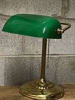 Front view of vintage bankers lamp showing green glass shade and solid brass base against concrete wall background.