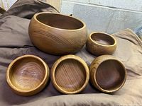 Five teak wooden bowls arranged with four smaller bowls in front and one larger bowl behind, showing wood grain and smooth finish
