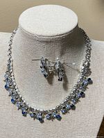 Front view of vintage Coro signed silver-tone necklace and matching earrings displayed on a cream fabric jewelry bust, showing alternating blue and clear rhinestones.