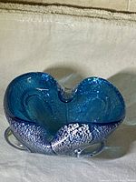 Top and angled side view of the blue glass ashtray showing the silver flakes embedded within the glass and the wavy organic shape with central notch.