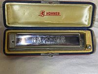 Open vintage case with fitted cream-colored padded interior and red Hohner logo, showing harmonica inside.