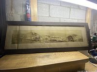 Full frontal view of the vintage panoramic black and white photograph inside an oak wooden frame, resting on wood surfaces, showing the wide shape and aged look.