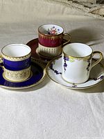 Three miniature tea cups with matching saucers: one blue and gold Aynsley, one pink floral Paragon, one white and purple floral with gold
