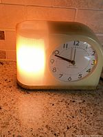 Front view of the clock showing the illuminated built-in light on the left and clear view of clock face with brand and model information.