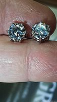 Close-up photo of two round cut moissanite stud earrings held on fingertips showing prong settings and sparkle.
