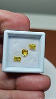 Three yellow sapphires (two rectangular, one heart-shaped) displayed in a small white jewelry box held between thumb and forefinger.