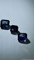 Three emerald cut blue sapphire gemstones laid out on neutral background, showing shape, color and clarity.